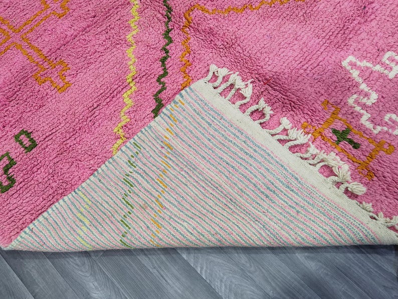 Op de afbeelding: Roze en wit geweven tapijt met een geometrisch patroon. Het tapijt heeft een franje rand en is gemaakt van zachte, pluche vezels.