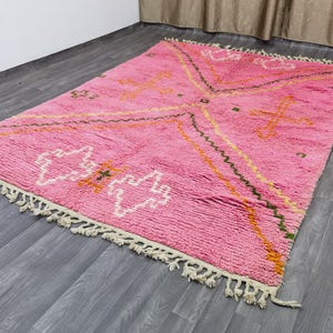 Op de afbeelding: Een roze en wit Marokkaans tapijt met een geometrisch patroon. Het tapijt heeft een franje rand en is gemaakt van wol.