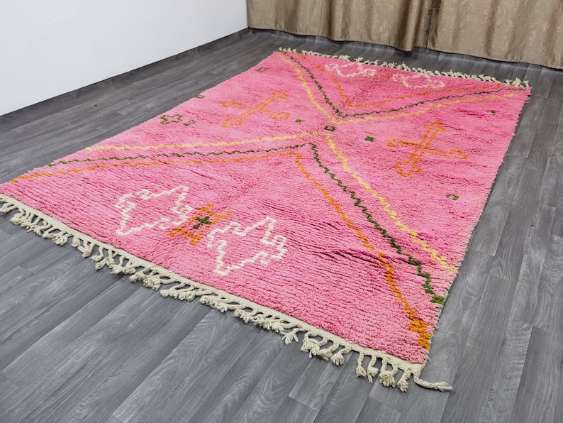 Op de afbeelding: Een roze en wit Marokkaans tapijt met een geometrisch patroon. Het tapijt heeft een franje rand en is gemaakt van wol.
