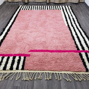 May include: A rectangular rug with a pink center and black and white striped borders. The rug has a fringe along the bottom edge and a hot pink accent stripe. The rug is made of a soft, textured material.