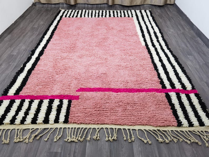 May include: A rectangular rug with a pink center and black and white striped borders. The rug has a fringe along the bottom edge and a hot pink accent stripe. The rug is made of a soft, textured material.