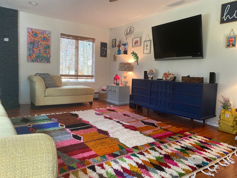 May include: A colorful, multi-patterned rug with a white background in a living room with a light blue dresser, a beige chaise lounge, and a large flat screen TV on the wall. The wall is decorated with artwork and the text "hello".