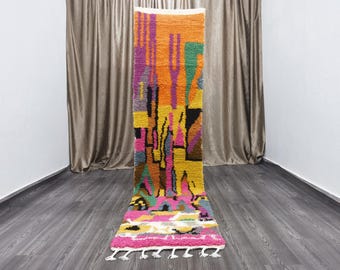 May include: A colorful abstract rug with geometric shapes in shades of pink, orange, yellow, green, and purple. The rug has a fringe border and is laid out on a gray wood floor.