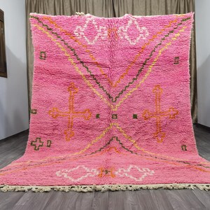 Op de afbeelding: Een roze Marokkaanse tapijt met een geometrisch patroon in tinten oranje, geel en groen. Het tapijt heeft een franje rand.