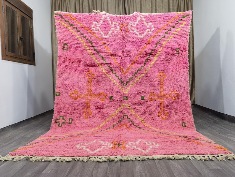 Op de afbeelding: Een roze Marokkaanse tapijt met een geometrisch patroon in tinten oranje, geel en groen. Het tapijt heeft een franje rand.