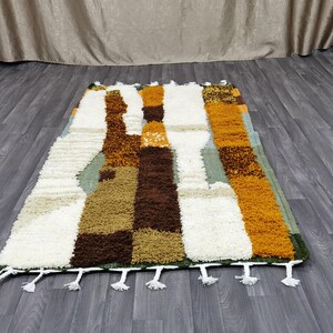 May include: Rectangular rug with a geometric pattern in shades of white, brown, tan, orange, and green. The rug has a shaggy texture and white tassels along the bottom edge. The rug is placed on a wooden floor.