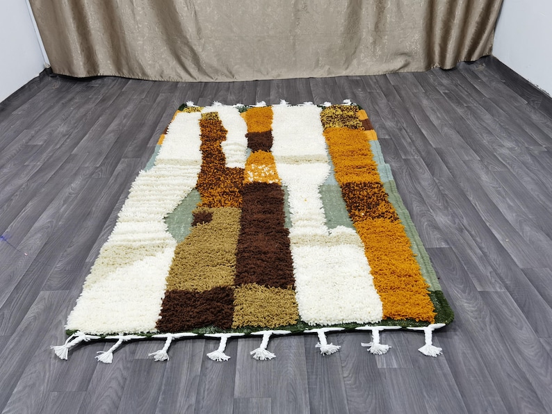 May include: Rectangular rug with a geometric pattern in shades of white, brown, tan, orange, and green. The rug has a shaggy texture and white tassels along the bottom edge. The rug is placed on a wooden floor.