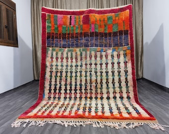 May include: A colorful Moroccan rug with a geometric pattern in shades of red, orange, blue, green, and white. The rug has a fringe border and is made of wool.