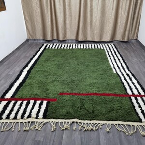 May include: A green rectangular rug with a geometric design. It has black and white striped borders, a red horizontal line, and cream fringe. The rug is on a wooden floor. The rug's dimensions are in centimetres.