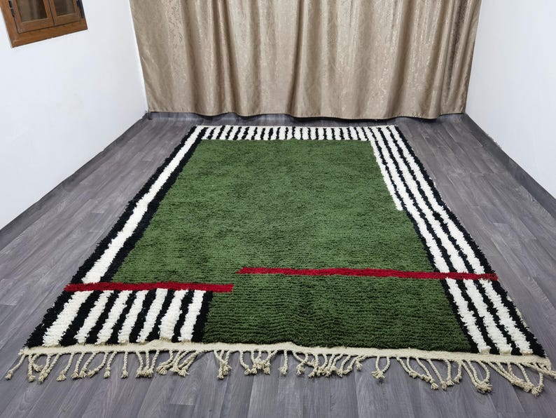 May include: A green rectangular rug with a geometric design. It has black and white striped borders, a red horizontal line, and cream fringe. The rug is on a wooden floor. The rug's dimensions are in centimetres.
