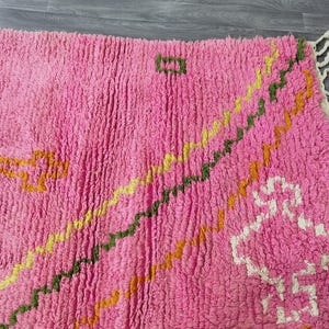 Op de afbeelding: Roze, gele en groene tapijt met franjes. Het tapijt heeft een pluche textuur en een geometrisch ontwerp.