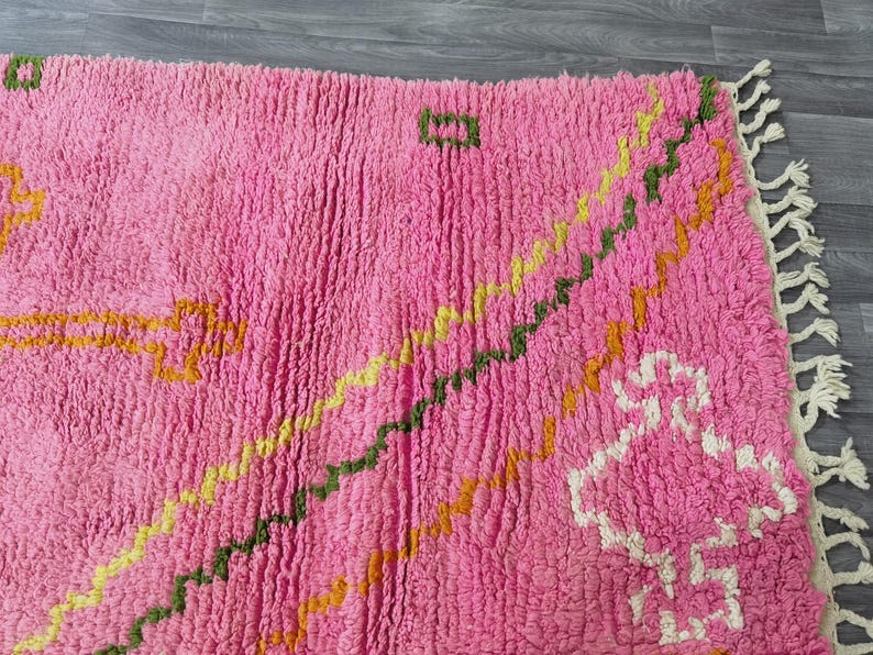 Op de afbeelding: Roze, gele en groene tapijt met franjes. Het tapijt heeft een pluche textuur en een geometrisch ontwerp.