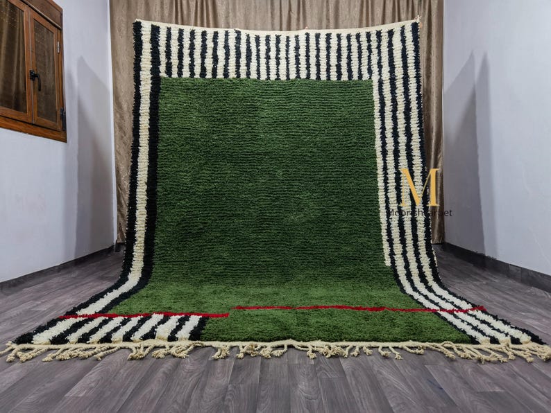 May include: A large, rectangular green rug with a textured surface. The rug features a border of alternating black and white vertical stripes. A red line accents the green field. The rug has a fringe along the bottom edge.