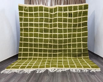 May include: A large, olive green rug with a grid pattern of white squares. The rug has a fringe along the bottom edge. The rug is made of a thick, woven material and is displayed against a neutral background.