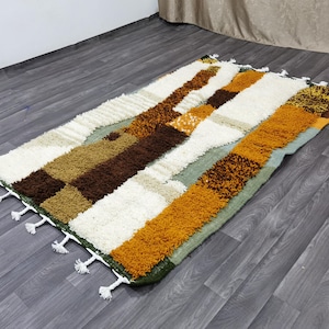 May include: Rectangular area rug with a patchwork design. The rug features a mix of colors including white, brown, tan, and orange. The rug has a shaggy texture and white tassels along the edges. The rug is placed on a wooden floor.