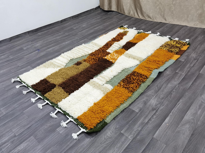 May include: Rectangular area rug with a patchwork design. The rug features a mix of colors including white, brown, tan, and orange. The rug has a shaggy texture and white tassels along the edges. The rug is placed on a wooden floor.