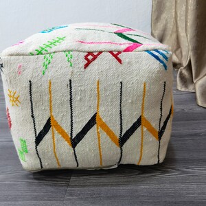 K&ouml;nnte beinhalten: Wei&szlig;er Pouf mit bunten geometrischen Mustern, darunter Schwarz, Gelb, Gr&uuml;n, Pink und Blau. Der Pouf ist aus gewebtem Stoff gefertigt und hat eine quadratische Form.