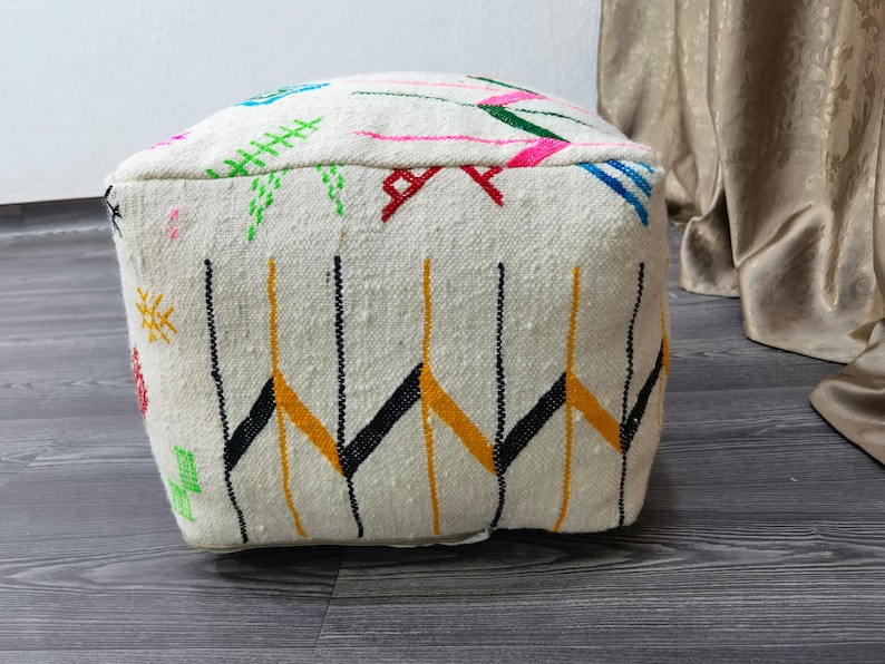 K&ouml;nnte beinhalten: Wei&szlig;er Pouf mit bunten geometrischen Mustern, darunter Schwarz, Gelb, Gr&uuml;n, Pink und Blau. Der Pouf ist aus gewebtem Stoff gefertigt und hat eine quadratische Form.