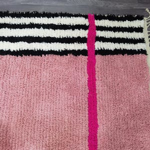 May include: A textured rug with a pink lower section and a black and white striped upper section. A bright pink vertical stripe runs down the center. The rug has a fringe on the right side.
