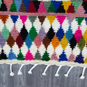May include: A colorful Moroccan rug with a diamond pattern and fringe. The rug is made of wool and has a variety of colors, including red, blue, green, yellow, and pink.