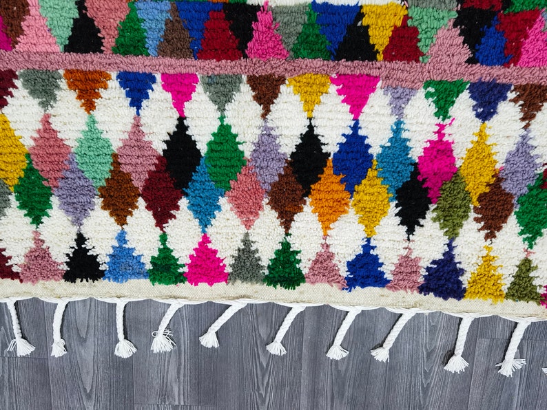May include: A colorful Moroccan rug with a diamond pattern and fringe. The rug is made of wool and has a variety of colors, including red, blue, green, yellow, and pink.