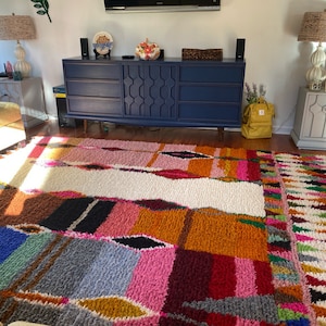 May include: A colorful, hand-woven rug with a geometric pattern in shades of blue, pink, orange, and white. The rug is made of wool and has a thick, plush texture.