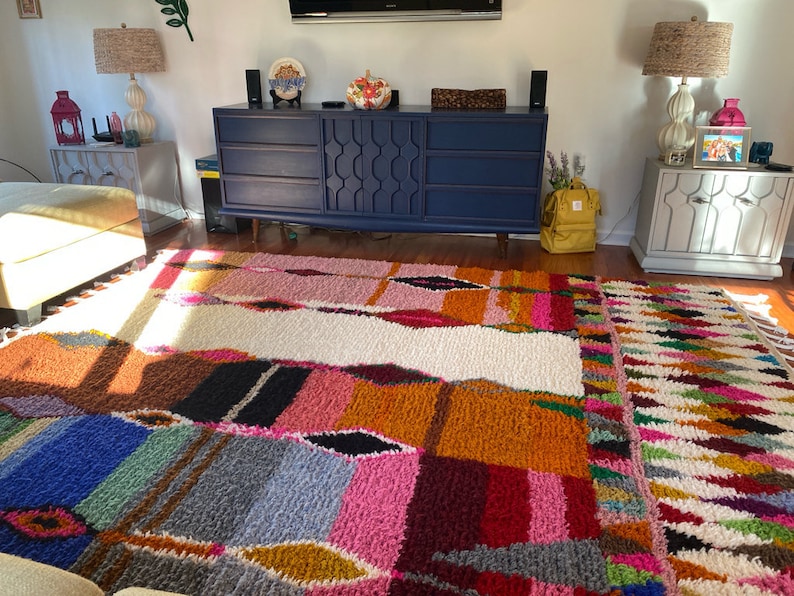 May include: A colorful, hand-woven rug with a geometric pattern in shades of blue, pink, orange, and white. The rug is made of wool and has a thick, plush texture.