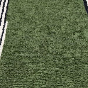 May include: A green rug with a textured surface. The rug has a border with black and white stripes on the left and right sides. The rug is made of a soft material.
