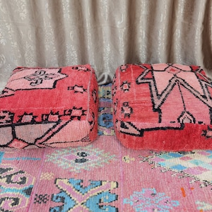 Op de afbeelding: Twee vierkante, rode Marokkaanse poefs met geometrische zwarte ontwerpen. De poefs staan op een roze tapijt met een kleurrijk patroon. De poefs zijn een levendig accent voor elke woonruimte.