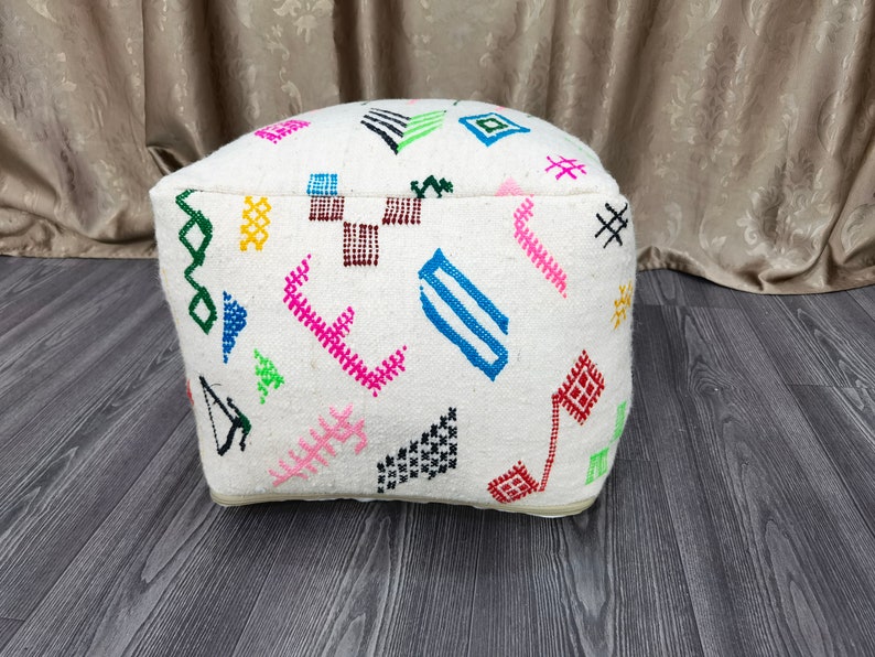K&ouml;nnte beinhalten: Wei&szlig;er Hocker mit bunten geometrischen Mustern, die auf den Stoff gestickt sind. Der Hocker ist quadratisch mit abgerundeten Kanten.
