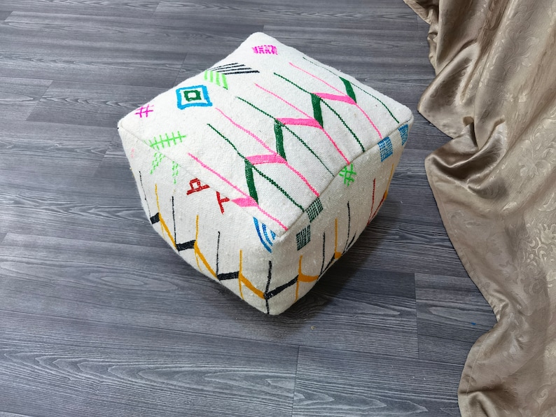 K&ouml;nnte beinhalten: Wei&szlig;er quadratischer Hocker mit bunten geometrischen Mustern in Rosa, Gr&uuml;n, Blau, Gelb und Schwarz. Der Hocker steht auf einem grauen Holzboden.