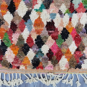 May include: A colorful Moroccan rug with a diamond pattern and fringe. The rug is made of wool and has a variety of colors, including red, orange, yellow, green, blue, and white.