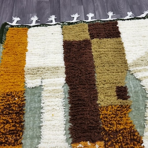 May include: A rectangular rug with a textured, shaggy pile. The rug features vertical stripes in shades of gold, brown, white, and olive green. The top edge has a dark green border with white tassels. This rug is suitable for home decor.