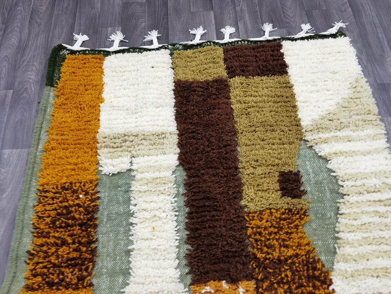 May include: A rectangular rug with a textured, shaggy pile. The rug features vertical stripes in shades of gold, brown, white, and olive green. The top edge has a dark green border with white tassels. This rug is suitable for home decor.