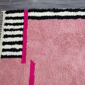May include: A close-up of a pink rug with a geometric design. The rug features a section of black and white stripes, a vertical fuchsia stripe, and a pink textured field. The rug has fringe on one edge.