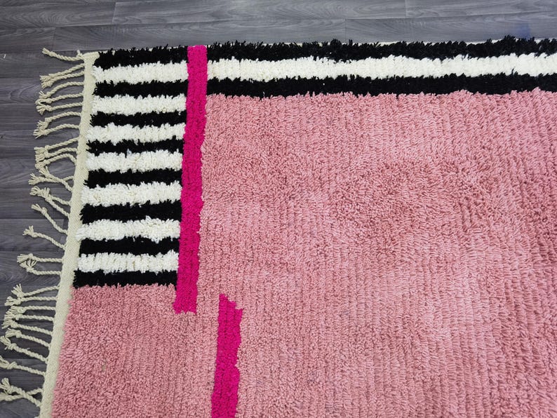 May include: A close-up of a pink rug with a geometric design. The rug features a section of black and white stripes, a vertical fuchsia stripe, and a pink textured field. The rug has fringe on one edge.
