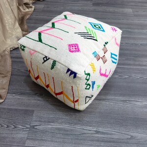 K&ouml;nnte beinhalten: Wei&szlig;er quadratischer Ottoman mit bunten geometrischen Mustern, die in den Stoff eingewebt sind. Der Ottoman steht auf einem grauen Holzboden.