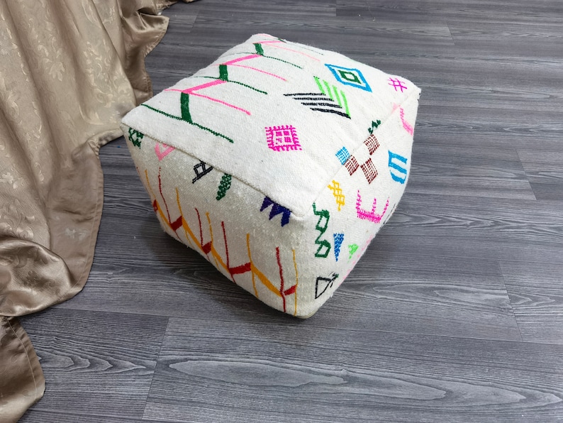 K&ouml;nnte beinhalten: Wei&szlig;er quadratischer Ottoman mit bunten geometrischen Mustern, die in den Stoff eingewebt sind. Der Ottoman steht auf einem grauen Holzboden.
