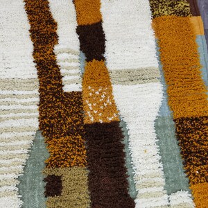 May include: A colorful, textured rug with a geometric pattern. The rug features vertical stripes in shades of white, brown, orange, and tan. The rug has a shaggy, plush texture, and is likely made of a soft material.