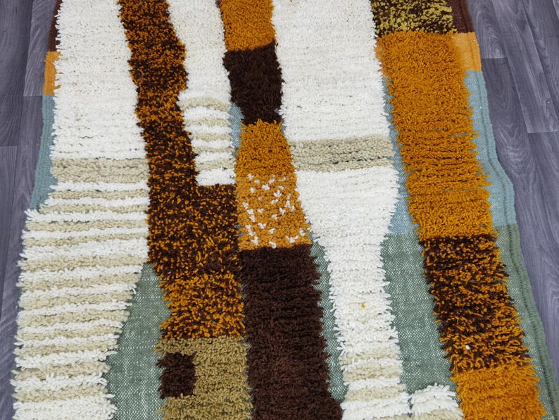 May include: A colorful, textured rug with a geometric pattern. The rug features vertical stripes in shades of white, brown, orange, and tan. The rug has a shaggy, plush texture, and is likely made of a soft material.