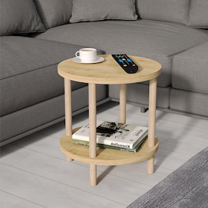 Könnte beinhalten: Ein Beistelltisch aus hellem Holz mit runder Tischplatte und einem unteren Regal. Der Tisch hat vier Beine und steht auf einem grauen Teppich. Eine Fernbedienung und eine Tasse Kaffee befinden sich auf dem oberen Regal.