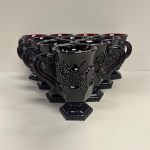 May include: A collection of ten black glass cups with ornate handles and bases. The cups have a dark red interior and are arranged in a semi-circular pattern. The cups have a detailed, embossed floral design.
