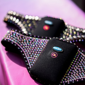 May include: Two black exercise belts with iridescent rhinestones and a blue button that says "TUNE BELT". The belts are on a pink surface.
