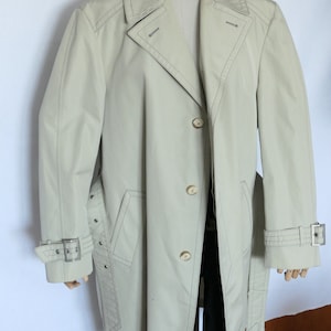 Puede incluir: Una gabardina beige con cinturón y cierre de botones. La gabardina tiene un frente de doble botonadura y un cuello.