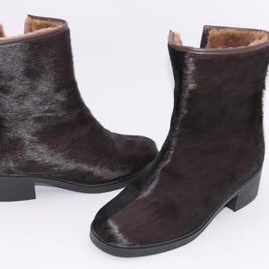 Könnte beinhalten: Ein Paar dunkelbraune Stiefeletten mit fellartiger Textur. Die Stiefel haben einen kleinen Absatz und ein weiches, hellbraunes Innenfutter. Die Stiefel sind vor einem weißen Hintergrund platziert.