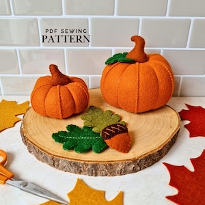 May include: A PDF sewing pattern featuring two orange felt pumpkins with brown stems, a green felt oak leaf, and a brown felt acorn. The items are displayed on a round wood slice, with additional felt maple leaves and orange scissors nearby.