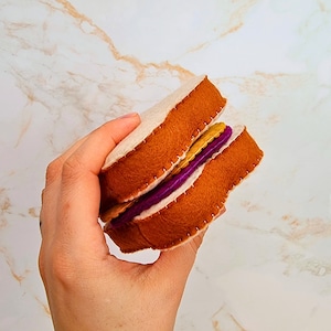 Może przedstawiać: Filcowa kanapka z brązowym chlebem, żółtym nadzieniem i fioletowym nadzieniem. Kanapka jest ręcznie szyta białą nicią. Chleb ma jasnobrązowy kolor.