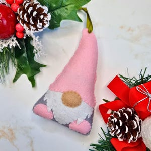 May include: A pink felt gnome ornament with a white beard, brown nose, and gray felt feet. The gnome is hanging from a green ribbon.