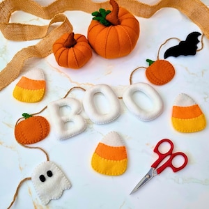 Op de afbeelding: Een verzameling vilten Halloween-decoraties met twee gevulde oranje pompoenen, drie snoepmaïs-vormen, een wit spookje, een zwarte vleermuis, een wit viltbanner met de tekst BOO, een rode schaar en een lint van jute.