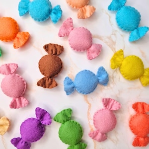 Könnte beinhalten: Eine Sammlung bunter Filz-Bonbon-Dekorationen in verschiedenen Farbtönen, darunter Blau, Rosa, Orange, Gelb, Grün, Braun und Lila. Jedes Bonbon hat eine runde Mitte und dekorative Seitenteile, angeordnet auf einer marmorierten Oberfläche.